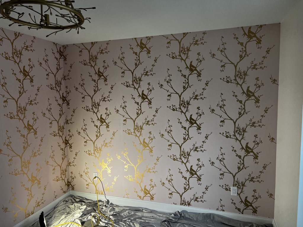 Bedroom with a gold bird-and-branch peel-and-stick wallpaper on pale walls, looking acceptable from across the room before the seams are seen up close.
