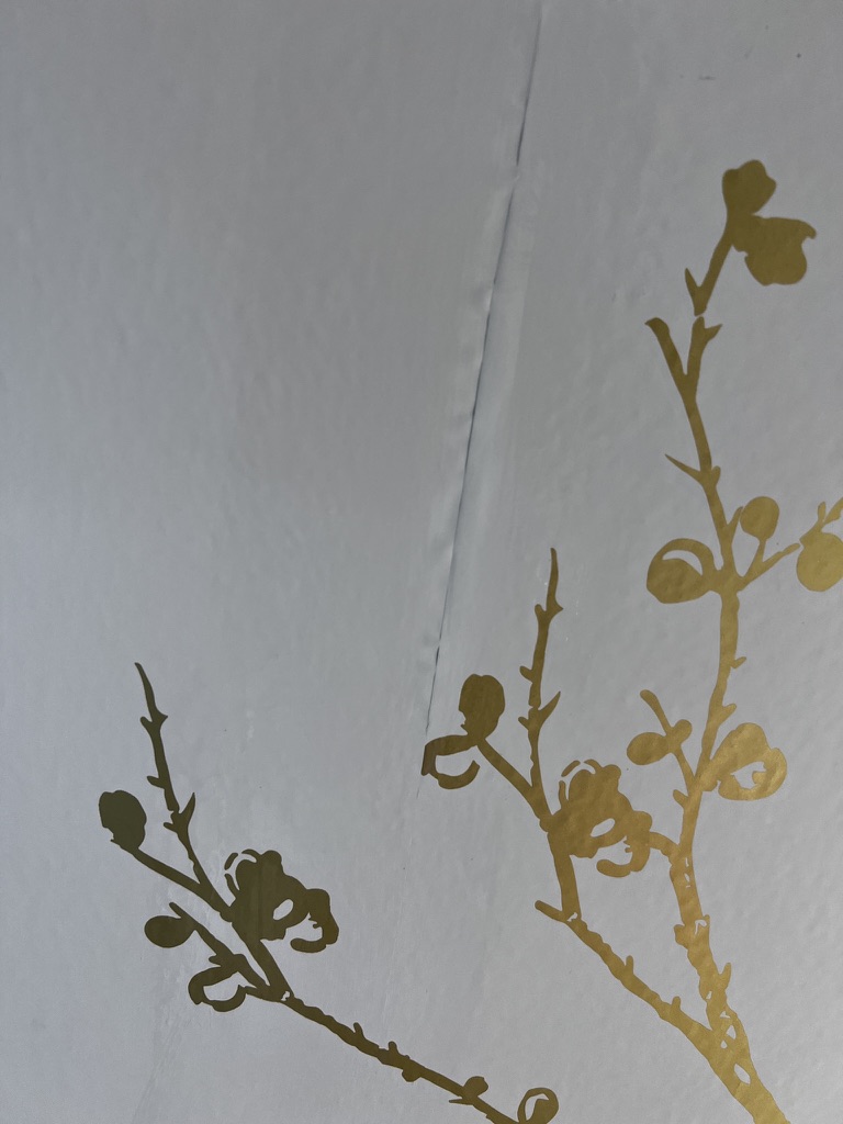 Peel-and-stick wallpaper pulling away over a taped-and-mudded drywall joint on a freshly painted new-construction wall.