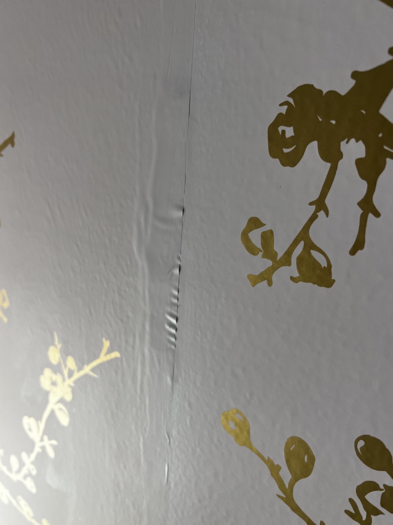 Peel-and-stick wallpaper puckering and ridging along a drywall mud joint, where fresh flat paint gave the adhesive nothing to bond to.