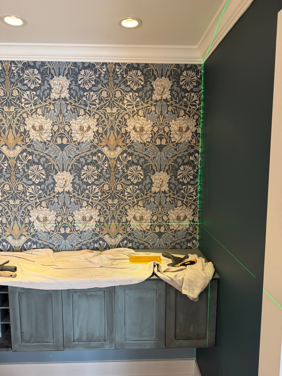 Green cross-line laser level projecting horizontal and vertical guides on a Morris & Co. Honeysuckle & Tulip focal wall during installation.
