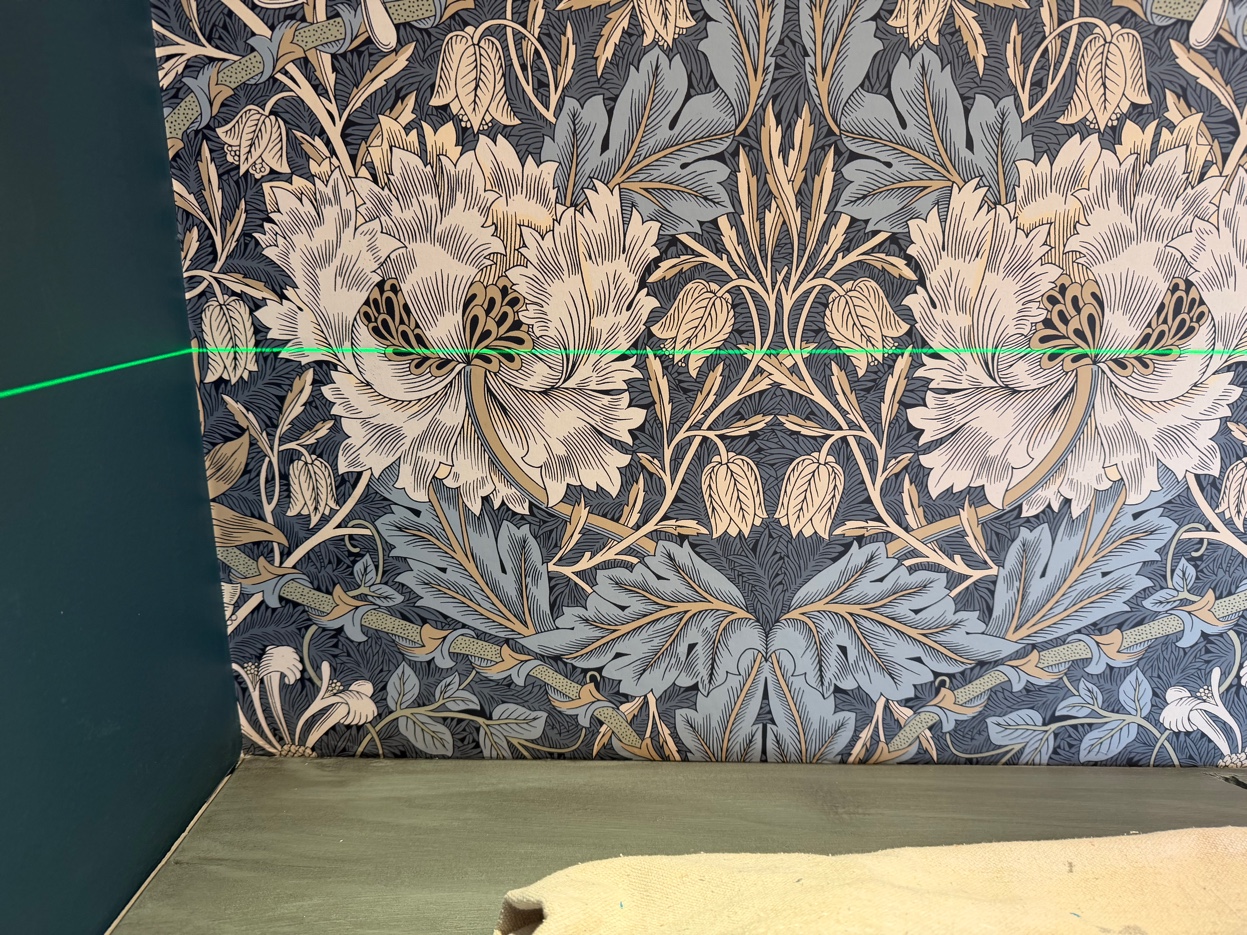 Seam and pattern-matching detail on Morris & Co. Honeysuckle & Tulip wallpaper near a wall corner, with a green laser reference line.