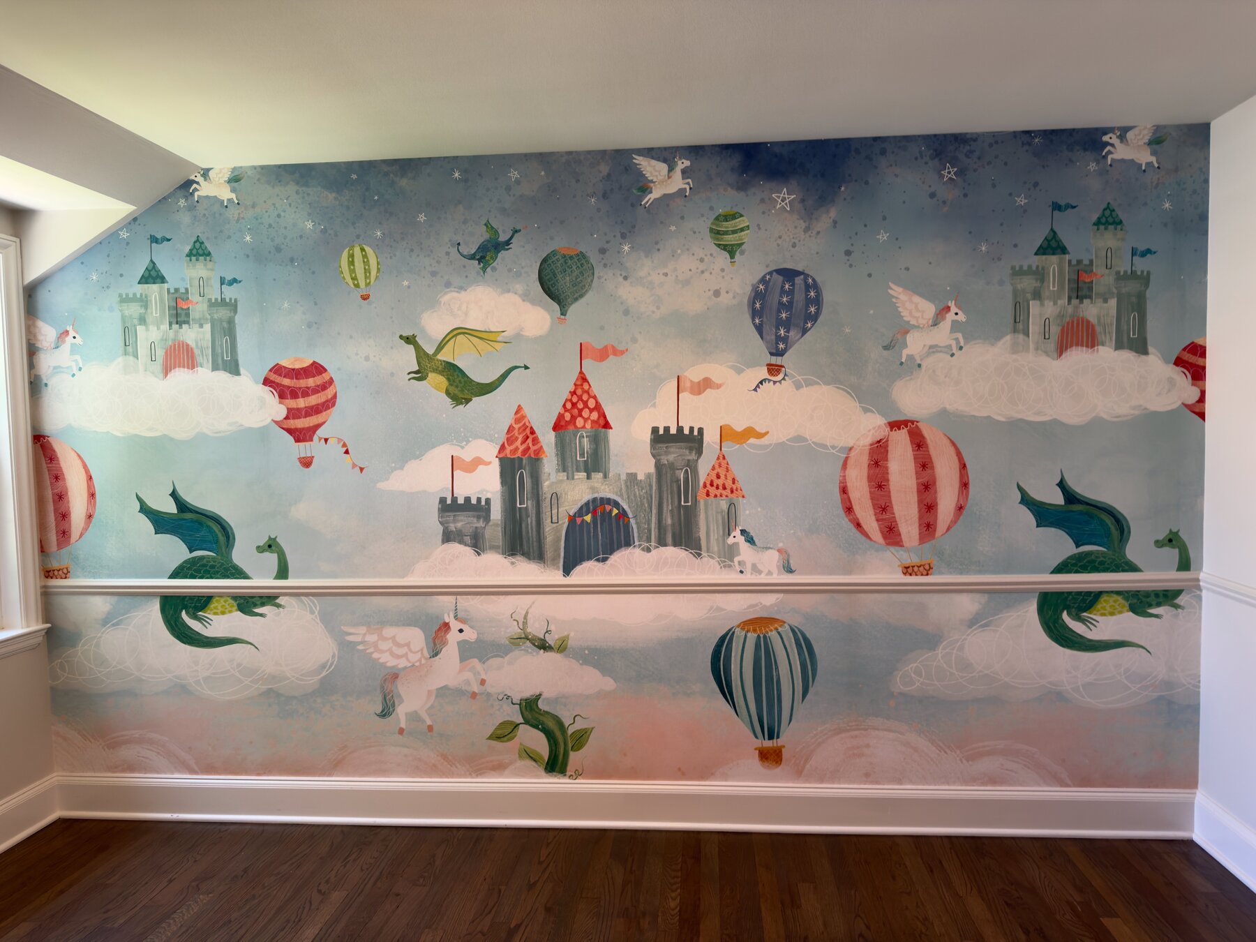 Finished nursery mural installation showing castles, dragons, unicorns, and hot air balloons wrapping a sloped ceiling