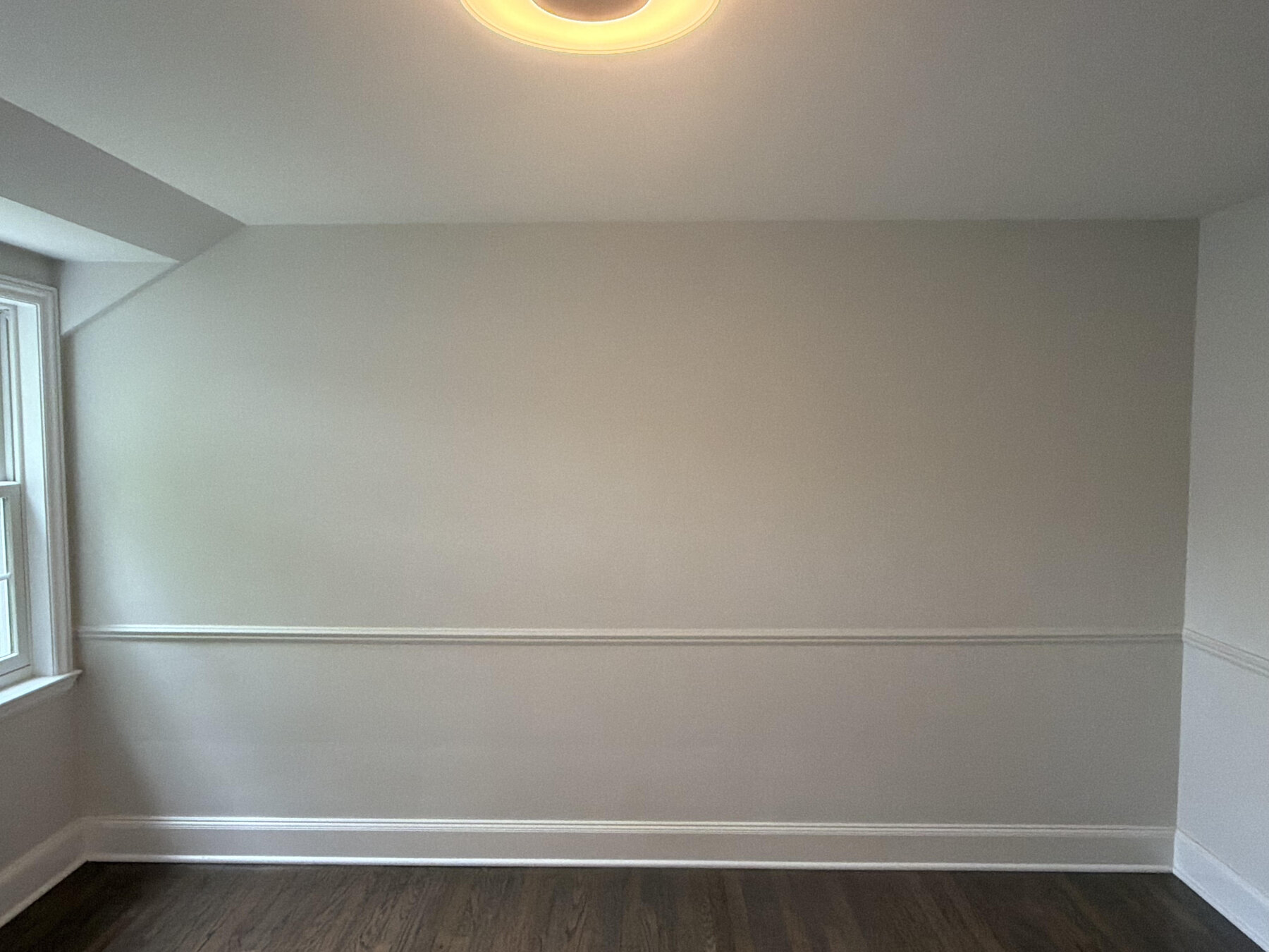 Bare nursery wall before mural installation, showing sloped ceiling, window, and chair rail