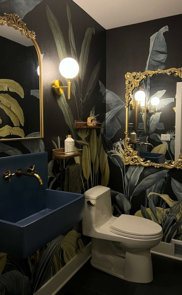 A dark powder room with a tropical leaf mural on a black ground, a gold globe sconce, an ornate gold mirror and a blue wall-mounted sink with gold taps.