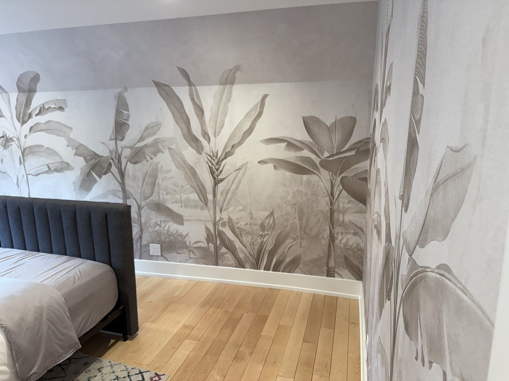 Rebel Walls banana leaf mural in a near-monochrome sepia palette, wrapping from the bed wall onto the return wall of a Michigan bedroom.