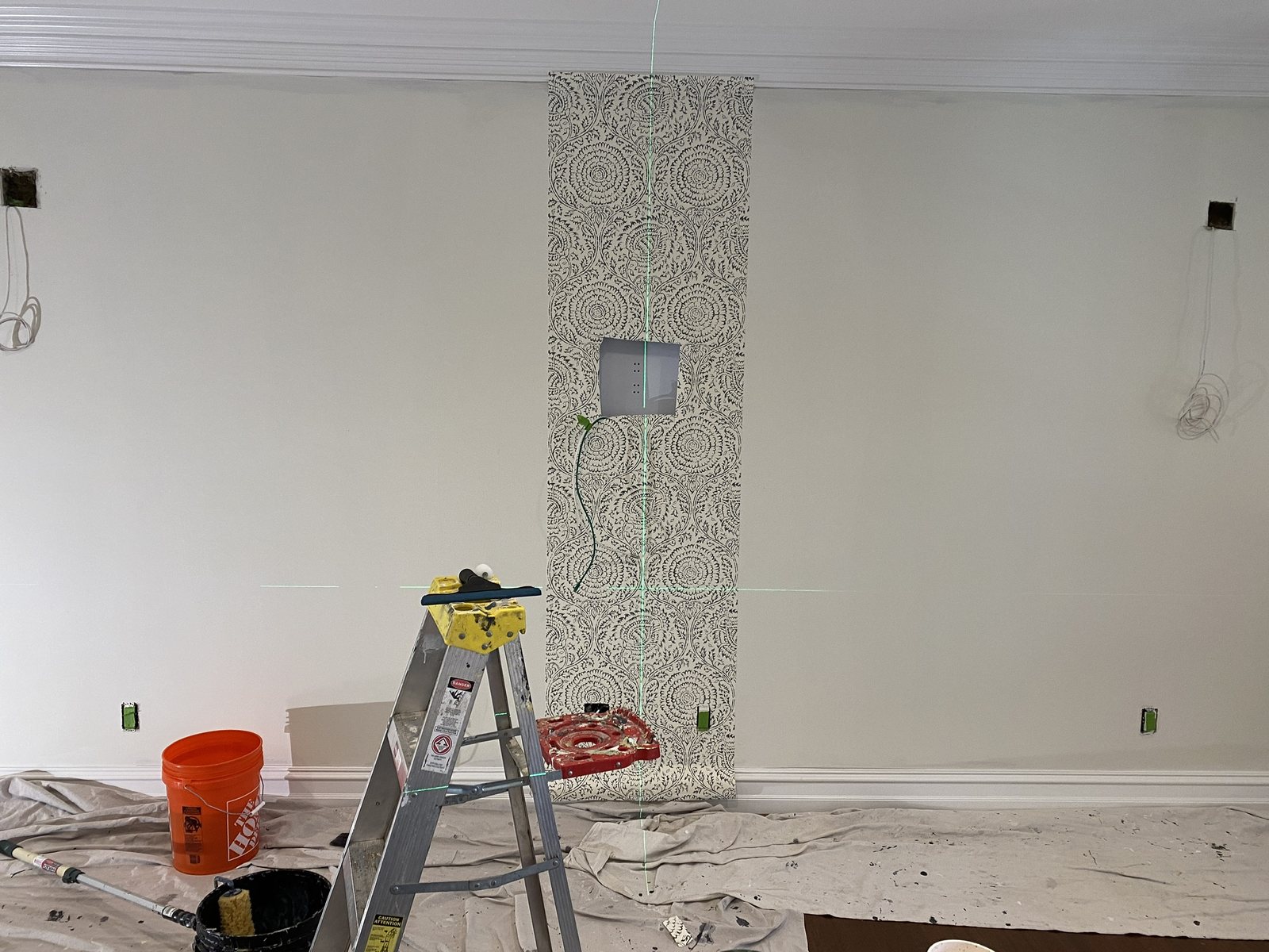The first strip of a black and cream medallion wallpaper hung against a laser layout line, with ladder, paste bucket and drop cloths in a Michigan room.