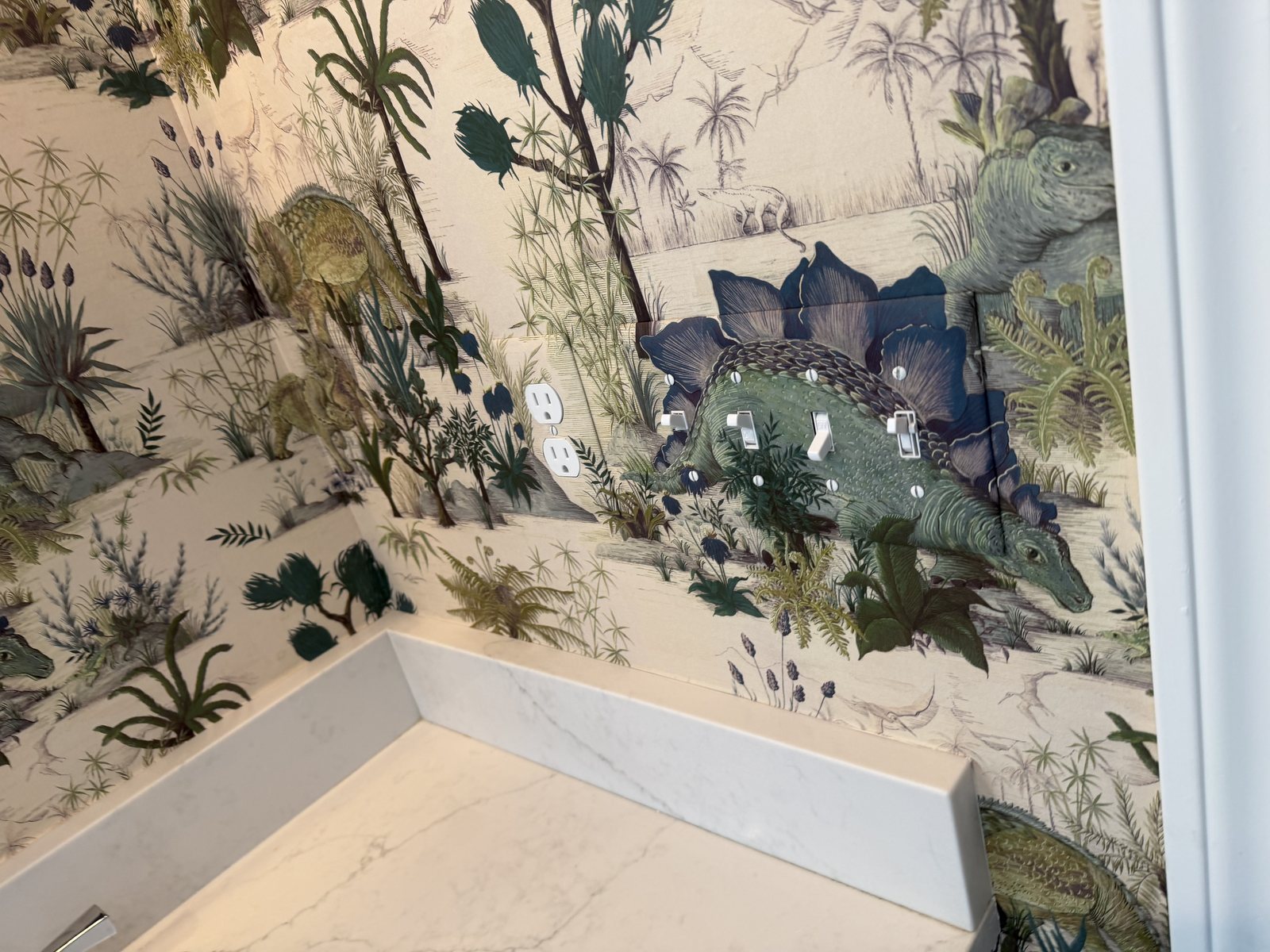 Dinosauria wallpaper panels turning an inside corner above the vanity backsplash, with the papered switch plates at right.
