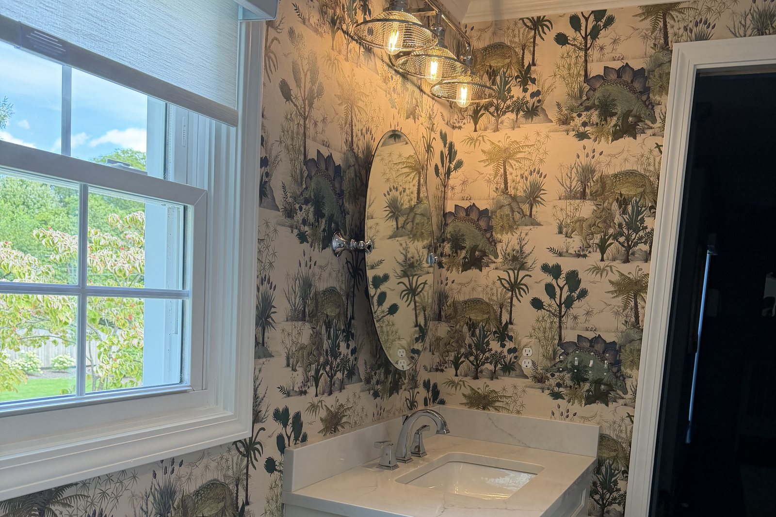 House of Hackney Dinosauria Ecru wallpaper in a children's bathroom in Birmingham, Michigan, with a white vanity, oval mirror and window.